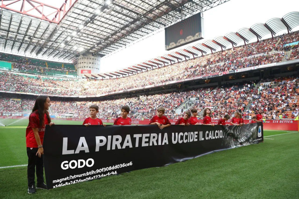 Foto in uno stadio, bambini mantengono uno striscione di propaganda TIM "LA PIRATERIA UCCIDE IL CALCIO.", l'immagine è stata alterata per includere alla fine la scritta "GODO"